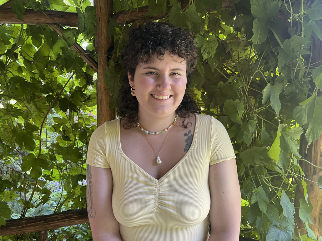 Picture of Kai. They have shoulder-length, dark-brown curly hair. They are wearing a pastel yellow shirt and are standing in front of a wall of leaves.
